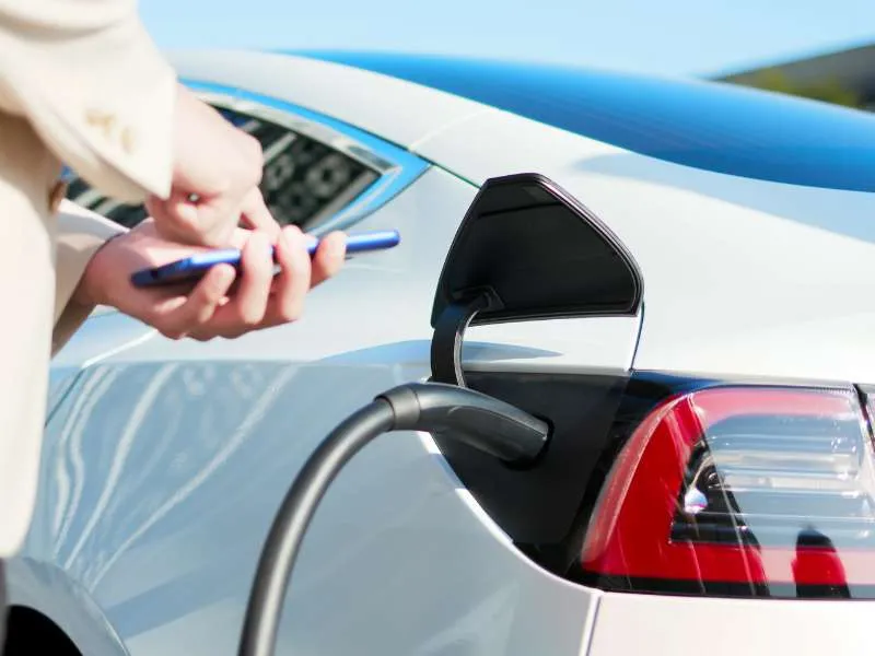 Električni automobil na punjaču, dok osoba u pozadini drži pametni telefon.